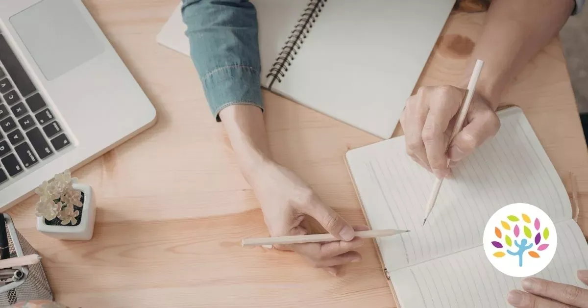 Hands writing in notebooks at a wooden desk with a laptop and coffee mug