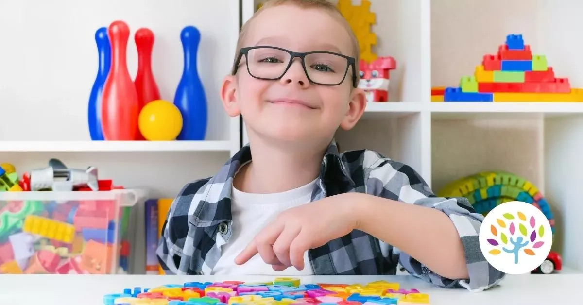 Smiling child with glasses at a colorful play table, with toys and blocks on shelves behind them