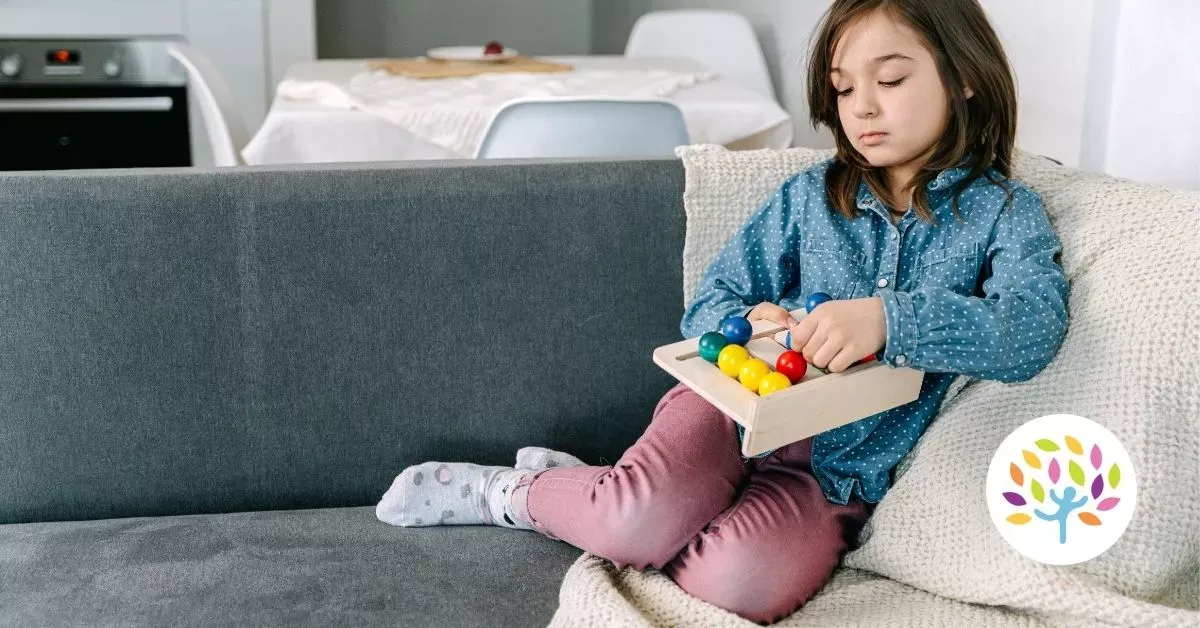 Child sitting on a couch, looking at a tray of colorful toys in a bright living room.