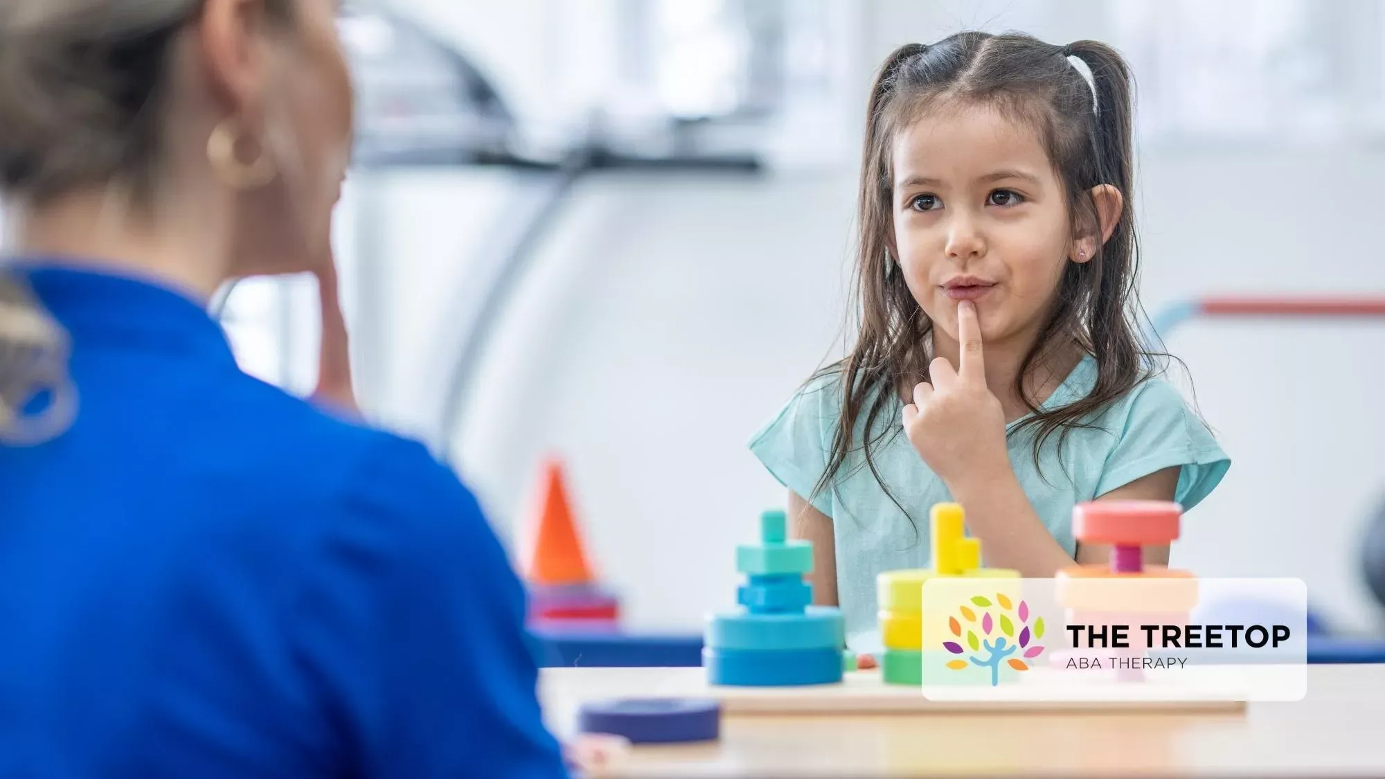 Child sitting at a therapy table, touching lips while looking at a clinician, with toys in front of her.