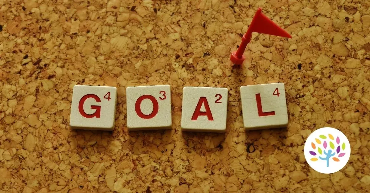 The word "GOAL" spelled out with Scrabble tiles on a cork board next to a small red flag pin.