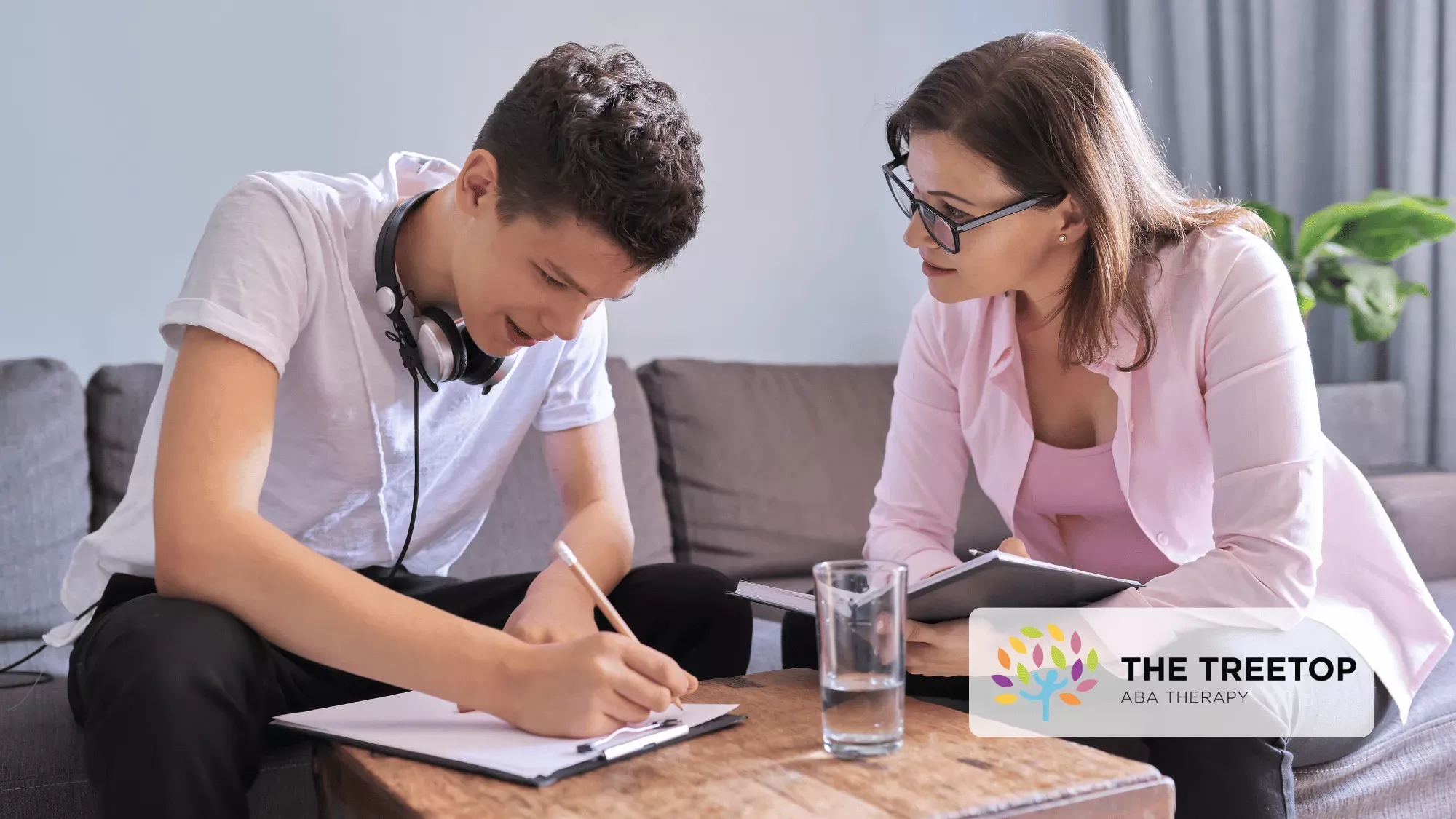 An adult and a teen work together at a table, one writing in a notebook while the other looks on.