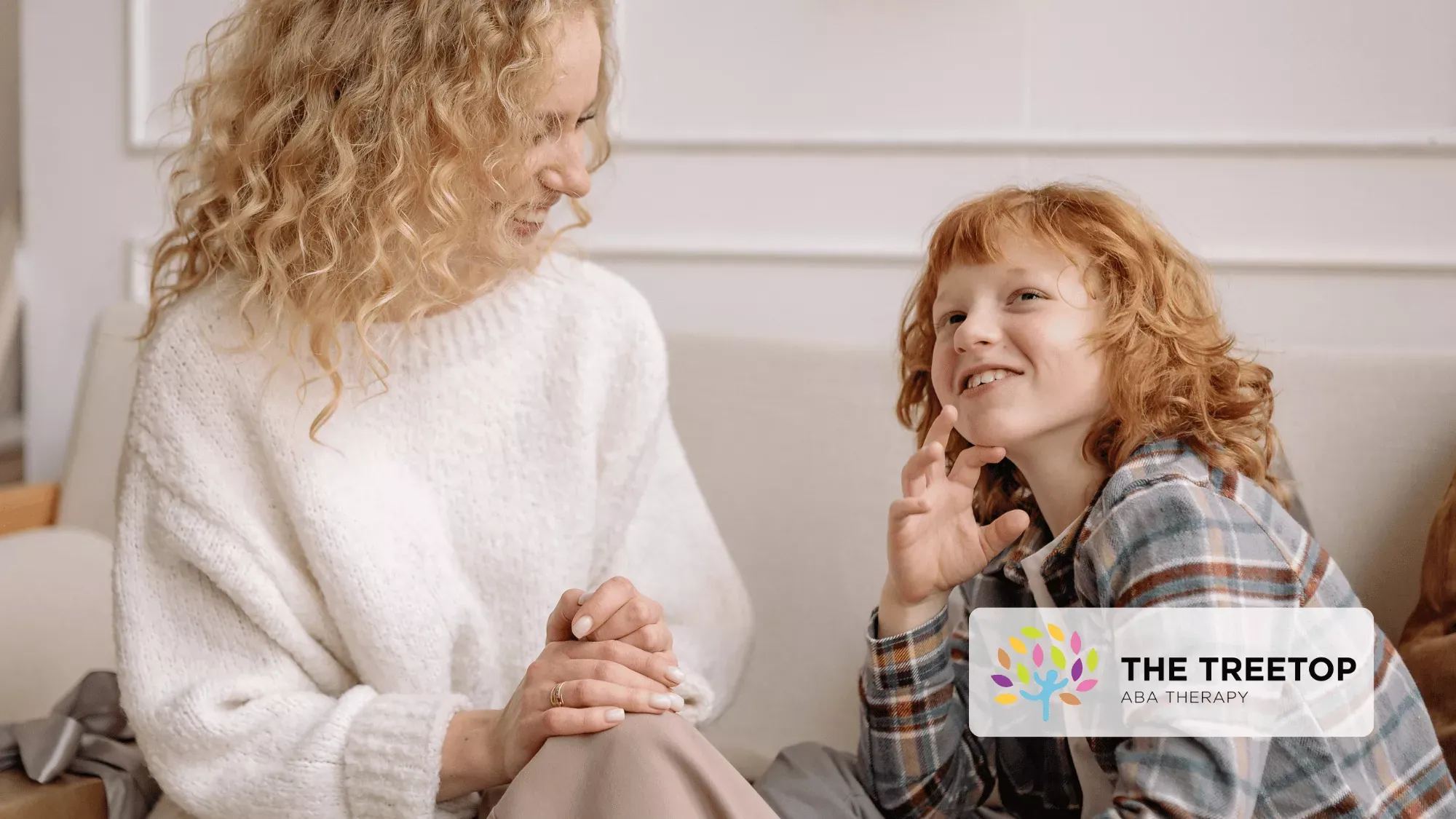 A person with curly hair smiling while talking to a child who is listening intently on a couch.