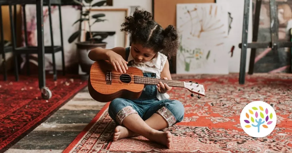 Child sitting on a patterned rug, strumming a small guitar indoors near houseplants.
