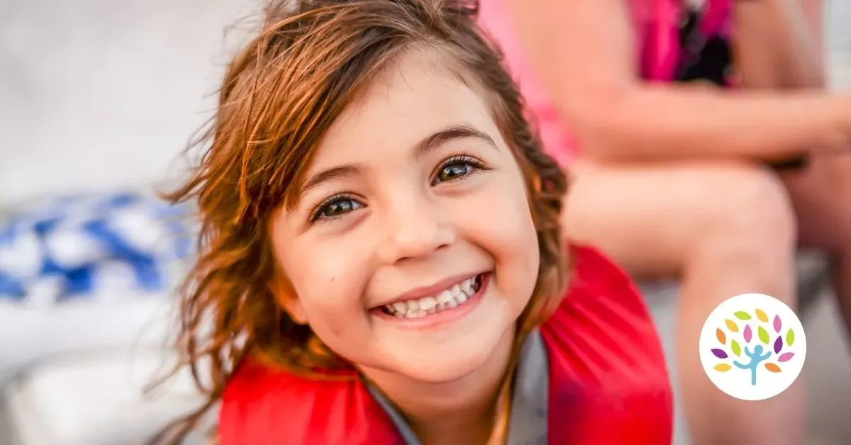 Smiling child with windblown hair in a red life vest, outdoors near water