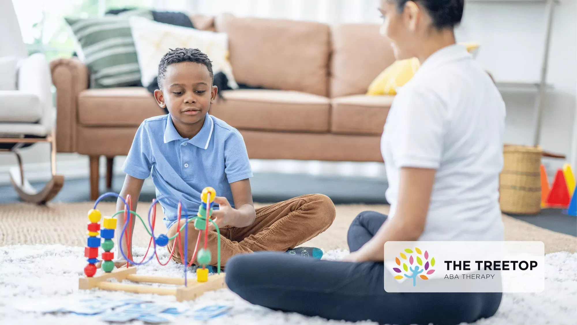 A child plays with a wooden bead maze on a white rug while sitting across from a professional in a living room.